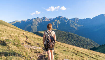 Back view anonymous female backpacker in casual wear walking on grassy lush hill slope and admiring picturesque scenery of severe mountain ridge on clear day - ADSF39019