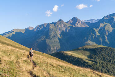 Back view anonymous female backpacker in casual wear walking on grassy lush hill slope and admiring picturesque scenery of severe mountain ridge on clear day - ADSF39024