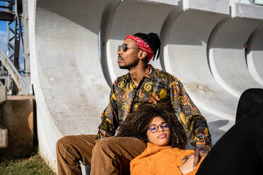 Black man sitting while woman with curly hair lies down in his lap looking away both wearing stylish outfits on concrete bench - ADSF39033