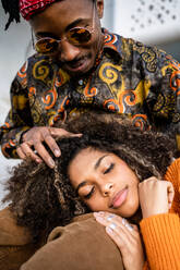 Glücklicher schwarzer Mann sitzt und streichelt das Haar der Frau, während sie in seinem Schoß mit geschlossenen Augen liegt und beide stilvolle Outfits auf einer Betonbank tragen - ADSF39036