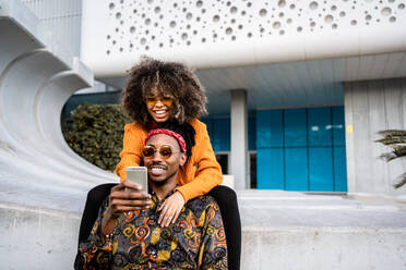 Moderner schwarzer Mann, der ein Mobiltelefon benutzt, mit charmanter ethnischer Frau mit Afrofrisur, die sich umarmt, während sie auf einer modernen Bank in der Stadt chillen - ADSF39038