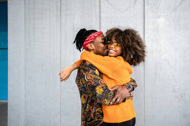 Seitenansicht liebender afroamerikanischer moderner Mann und Frau, die sich umarmen und fröhlich lächelnd gegen eine graue Wand auf der Straße stehen - ADSF39043