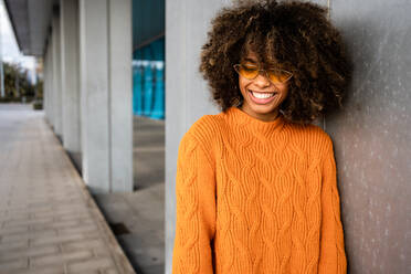 Lächelnde ethnische Frau mit lockigem Afro-Haar, die einen leuchtend orangefarbenen Pullover mit geschlossenen Augen trägt und sich gegen eine graue Wand lehnt - ADSF39049