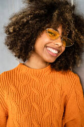 Beautiful cheerful ethnic woman with curly afro hair wearing vivid orange sweater looking at camera against gray wall - ADSF39052