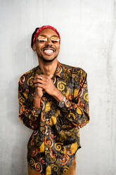 Glücklicher afroamerikanischer Mann mit Dreadlocks, der eine stilvolle runde Sonnenbrille mit rotem Stirnband und buntem Hemd trägt und lächelnd in die Kamera schaut - ADSF39056