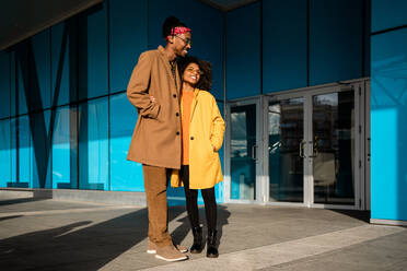 Ganzkörperaufnahme eines fröhlichen afroamerikanischen Mannes und einer Frau in schicken beigen und gelben Mänteln, die sich umarmen, während sie auf der Straße spazieren gehen und wegschauen - ADSF39059