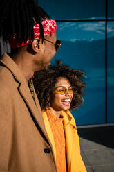 Cheerful African American man and woman in stylish beige and yellow coats embracing each other while walking on the on street with closed eyes - ADSF39060