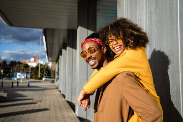 Cooler afroamerikanischer Mann mit Sonnenbrille und Mantel, der seine lachende Freundin mit Afro-Lockenhaar huckepack nimmt und in die Kamera schaut, während er Zeit auf der Straße verbringt - ADSF39065