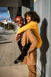 Cooler afroamerikanischer Mann mit Sonnenbrille und Mantel, der seine lachende Freundin mit Afro-Lockenhaar auf der Straße huckepack nimmt - ADSF39066