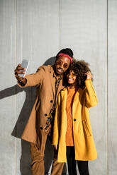 Cheerful black woman with curly hair embracing cool ethnic guy in sunglasses and smiling while taking selfie against gray wall in city - ADSF39068