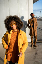 Thoughtful black woman with afro curly hair wearing vivid coat standing looking away on street with cool blurred guy on background - ADSF39084