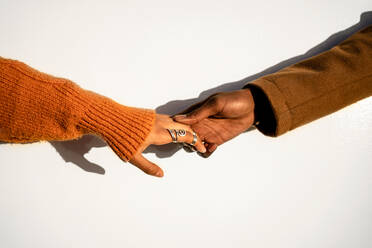 Crop woman in orange sweater and rings holding hand of black boyfriend in brown coat against wall in sunset light - ADSF39097