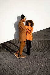 Cool African American man with dreadlocks wearing sunglasses with jacket standing at white wall close to stylish girlfriend with curly hair and closed eyes in sunset light - ADSF39103