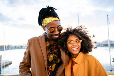 Strahlend lachende schwarze Frau in gelbem Mantel mit trendigem Freund mit Sonnenbrille, die eng beieinander stehen und wegschauen, auf einem städtischen Pier - ADSF39113
