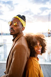 Seitenansicht einer fröhlich lachenden schwarzen Frau in gelbem Mantel mit einem trendigen Freund mit Sonnenbrille, der Rücken an Rücken steht und in die Kamera schaut, auf einem städtischen Pier - ADSF39117