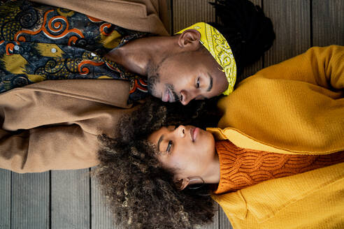 Top view of trendy African American man and woman in bright coat lying opposite with face to face looking at each other - ADSF39124