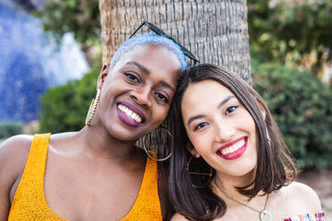 Delighted multiracial couple of lesbian women standing together near palm tree in city - ADSF39129