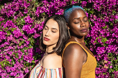 Seitenansicht von asiatischen und afroamerikanischen Freundinnen, die Rücken an Rücken neben einem hell blühenden Strauch im Garten stehen - ADSF39157