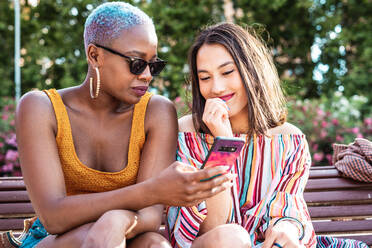 Multiracial lesbian couple sitting on bench and browsing smartphone together on blurred background of park on sunny day - ADSF39160