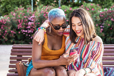 Multiracial lesbisches Paar sitzt auf der Bank und Surfen Smartphone zusammen auf unscharfen Hintergrund des Parks am sonnigen Tag - ADSF39161