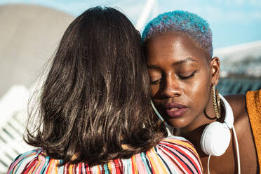 African American female with piercing touching hair of beautiful Asian girlfriend looking away on blurred background - ADSF39176