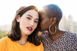 Afroamerikanische Frau mit Piercing, die das Haar ihrer schönen asiatischen Freundin berührt und in die Kamera schaut, auf unscharfem Hintergrund - ADSF39190