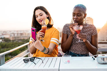 Multiracial girlfriends in trendy sunglasses sitting at table taking selfie on urban background - ADSF39213