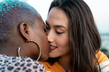 Junge multiethnische lesbische Frauen sitzen zusammen und küssen sich vor einem verschwommenen städtischen Hintergrund - ADSF39221