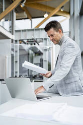 Lächelnder Geschäftsmann hält Papier und benutzt einen Laptop am Schreibtisch in der Industrie - DIGF18901