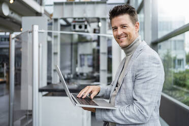 Smiling mature businessman holding laptop in industry - DIGF18906