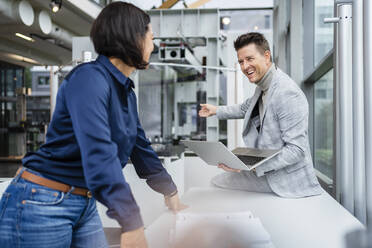 Happy businessman holding laptop talking with businesswoman at desk in industry - DIGF18909