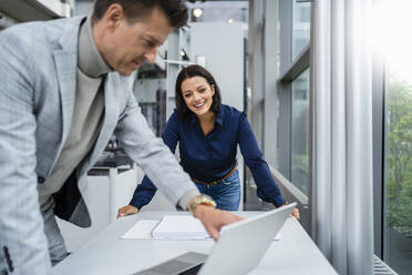 Happy businessman showing laptop to happy businesswoman at desk - DIGF18916