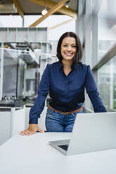 Smiling mature businesswoman with laptop at desk in industry - DIGF18922