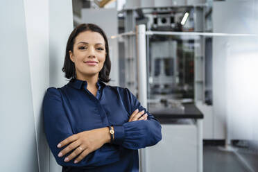 Smiling mature businesswoman with arms crossed leaning on wall in industry - DIGF18942