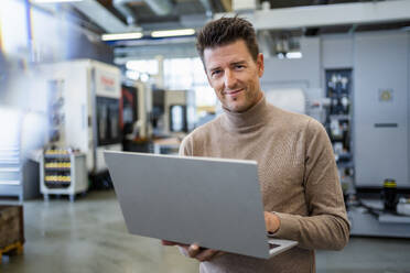 Lächelnder Geschäftsmann mit Laptop in der Industrie - DIGF18948