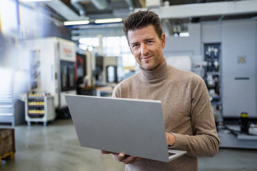 Smiling businessman holding laptop in industry - DIGF18948