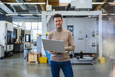 Lächelnder reifer Geschäftsmann mit Laptop in der Industrie - DIGF18950