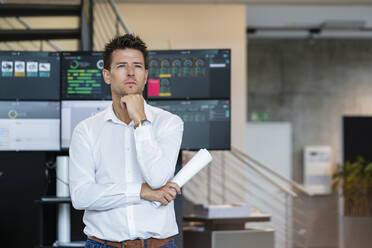 Nachdenklicher Geschäftsmann, der ein zusammengerolltes Papier hält und mit der Hand am Kinn vor einem Computerbildschirm steht - DIGF18974