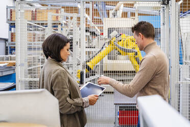 Businessman discussing with businesswoman holding tablet PC in front of robotic arm - DIGF18995