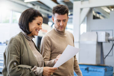 Happy businesswoman showing paper to businessman in production hall - DIGF19013