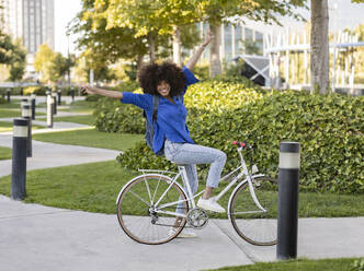 Happy Afro woman with arms raised sitting on bicycle at park - JCCMF07356