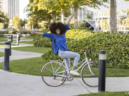 Glückliche Afro-Frau mit erhobenen Armen auf dem Fahrrad sitzend im Park - JCCMF07356