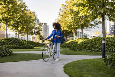 Lächelnde Frau mit Handy in der Hand, die mit dem Fahrrad im Park spazieren geht - JCCMF07357