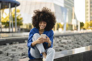 Lächelnde Afro-Frau mit Smartphone an der Wand sitzend - JCCMF07376