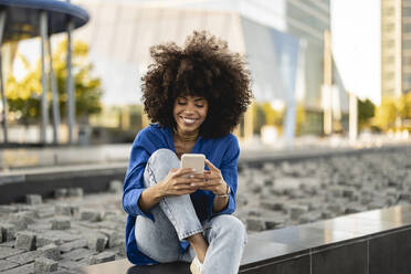 Smiling Afro woman using smart phone sitting on wall - JCCMF07376
