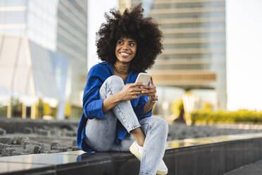 Smiling Afro woman holding smart phone sitting on wall - JCCMF07377