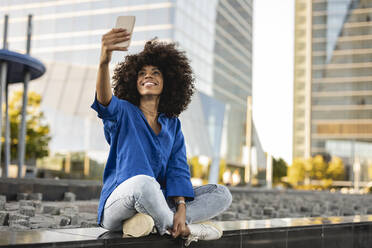 Glückliche Afro-Frau, die ein Selfie über ihr Handy macht und an der Wand sitzt - JCCMF07378