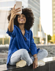 Glückliche Afro-Frau, die im Schneidersitz an der Wand sitzend ein Selfie durch ihr Smartphone macht - JCCMF07379