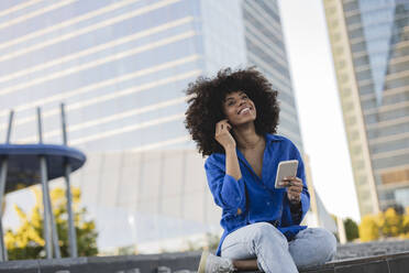 Lächelnde Frau mit Afrofrisur, die ein Mobiltelefon hält und an der Wand sitzt - JCCMF07380