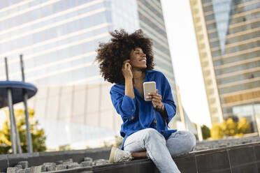Glückliche Afro-Frau mit Mobiltelefon an der Wand sitzend - JCCMF07381